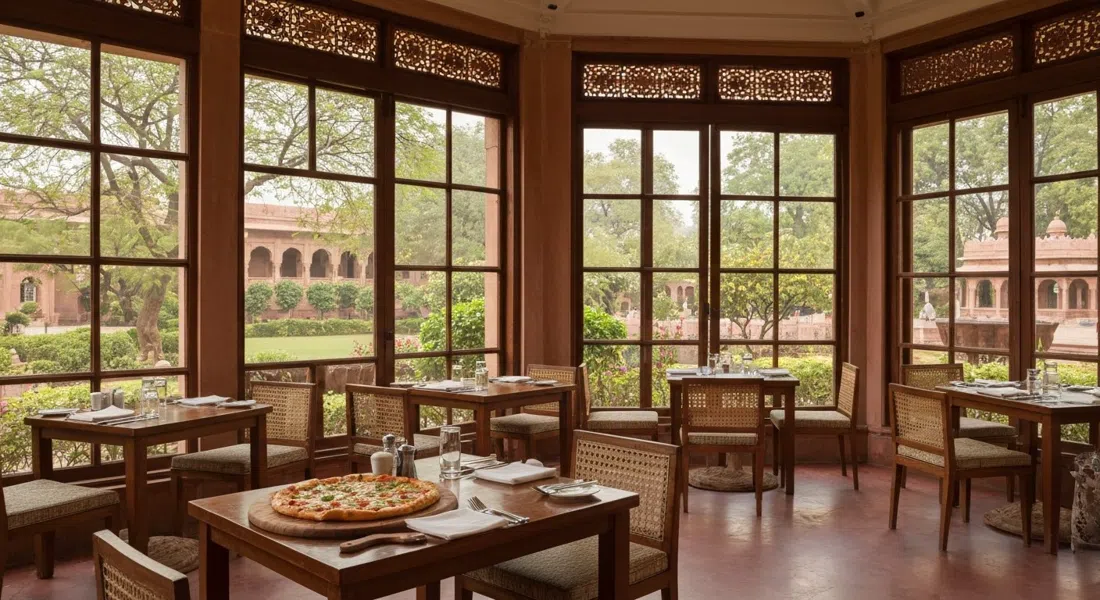 El comedor con vista al jardín del Trident Agra, pizza de horno de leña y jardines del hotel más allá de las ventanas bajo la suave luz de la tarde
